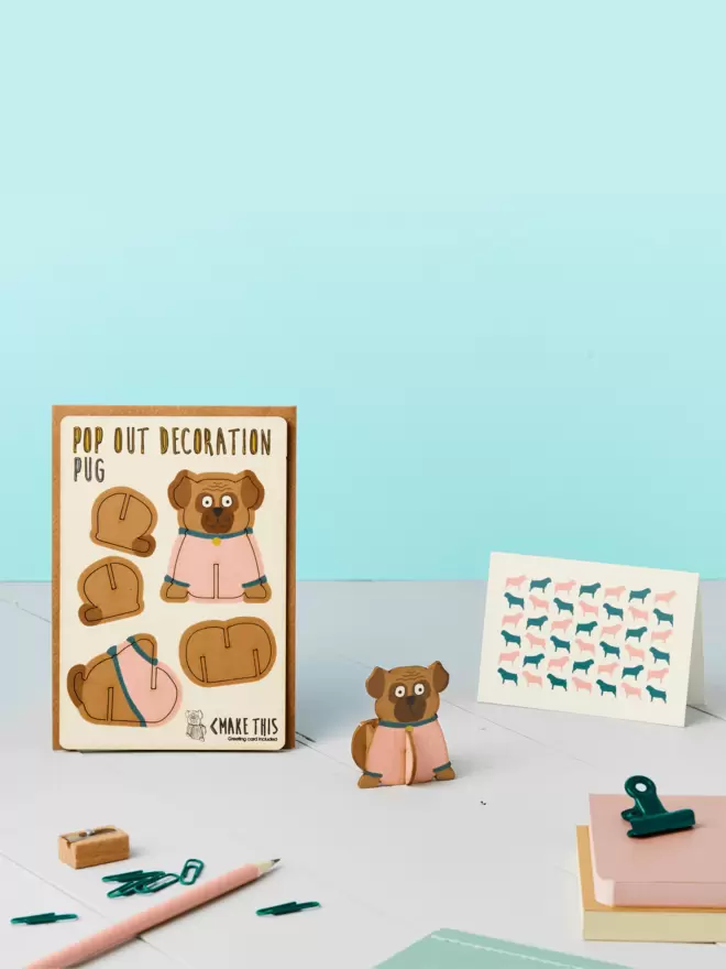 3D pug decoration and pug pattern greeting card and brown kraft envelope on top of a grey desk in front of a light blue coloured background