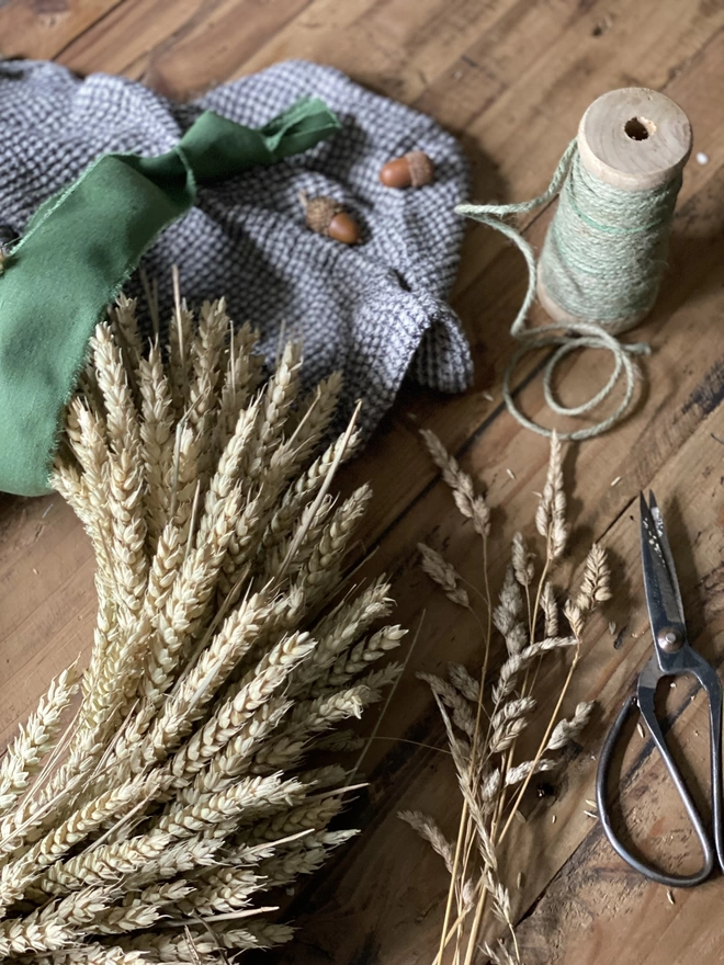 A close-up shot of the right curve of a handmade September Harvest Wheat Wreath with a sage green ribbon looped around the top, on display with a blue chequered cloth, small autumnal accessories and a few strands of loose wheat