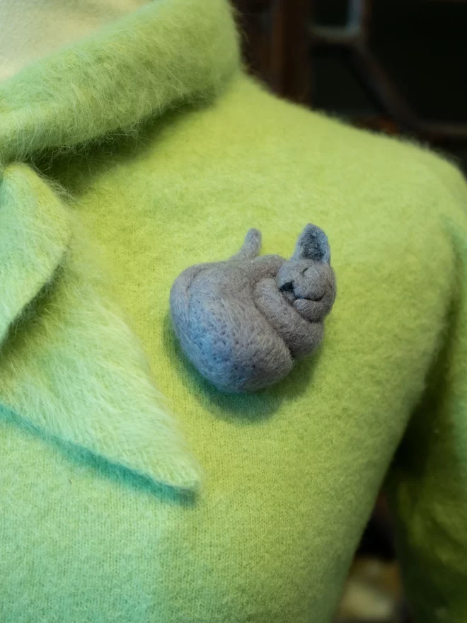 A needle felted grey sleeping cat brooch on a green fluffy jumper 