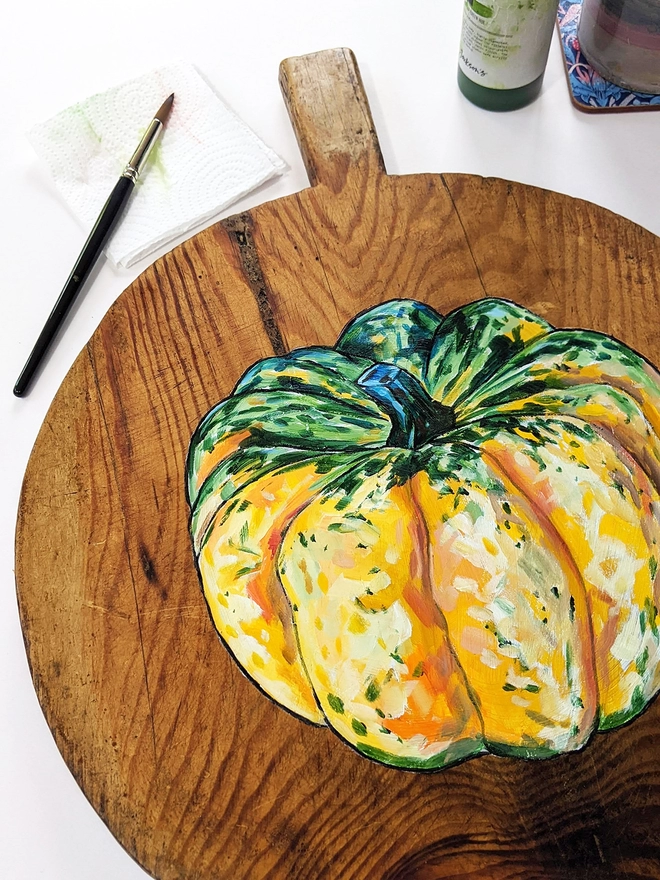 Wooden chopping board with carnival squash painted design on a table next to a paintbrush and paints