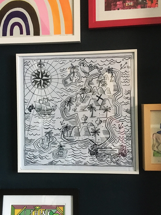 A Mr.PS Treasure map hankie shown in a picture frame on a dark wall surrounded by other framed prints