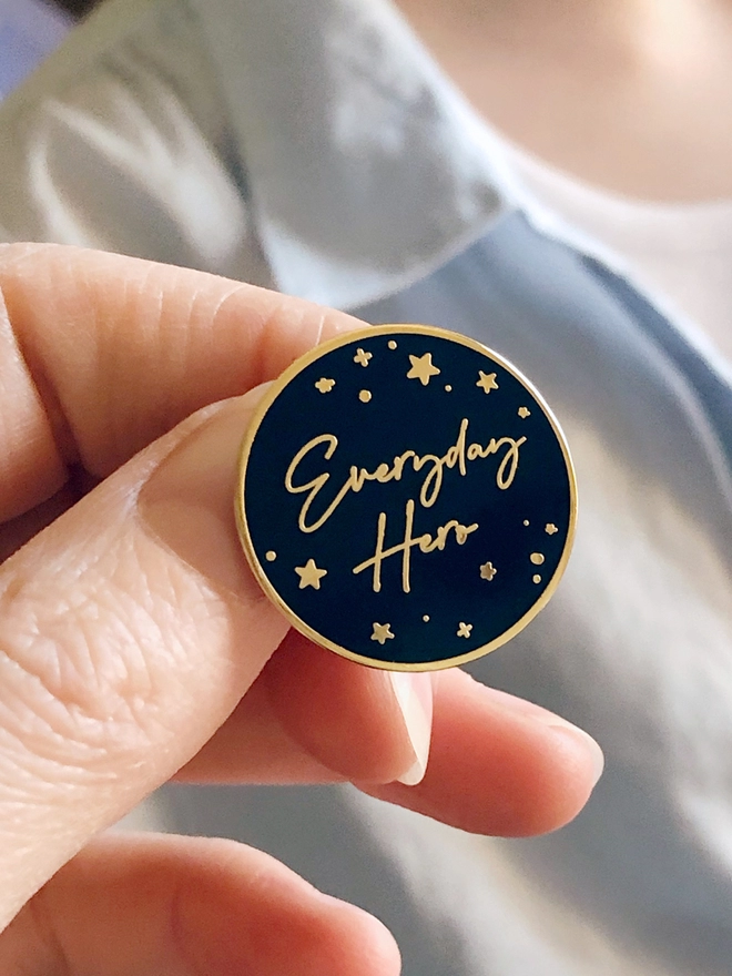 A hand is holding a navy blue and gold enamel pin badge with a gold star design and the words "Everyday Hero".