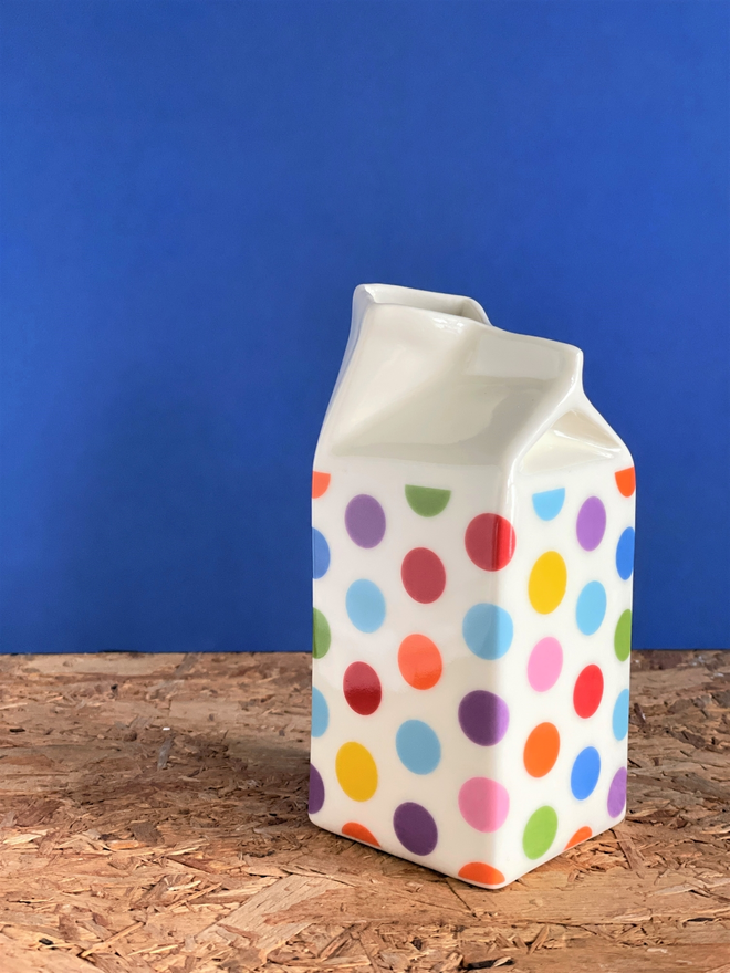 1 milk carton jug on a wooden surface  , decorated with multicoloured polkadot pattern in red , blue , green , orange , purple and yellow dots, the background is cobalt blue