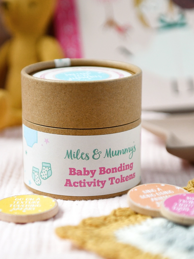 A cardboard jar with a label that reads Baby Bonding Activity Tokens stands on soft muslin fabric, with several wooden tokens beside it.