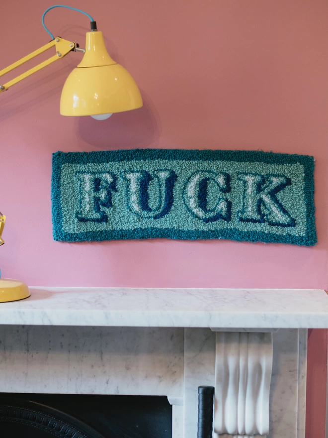 Blue 'FUCK' Handmade Tufted Rug/Wall Hanging seen on a pink wall above a mantlepiece with a yellow lamp.