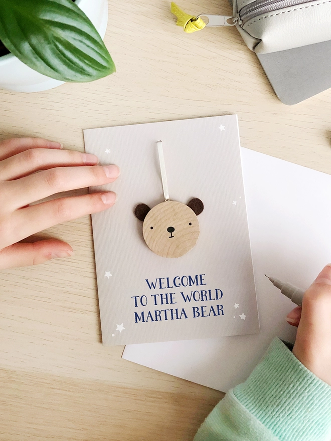 A grey greetings card with a small wooden bear keepsake and the words "Welcome to the world Martha bear" printed on is on a wooden desk. 