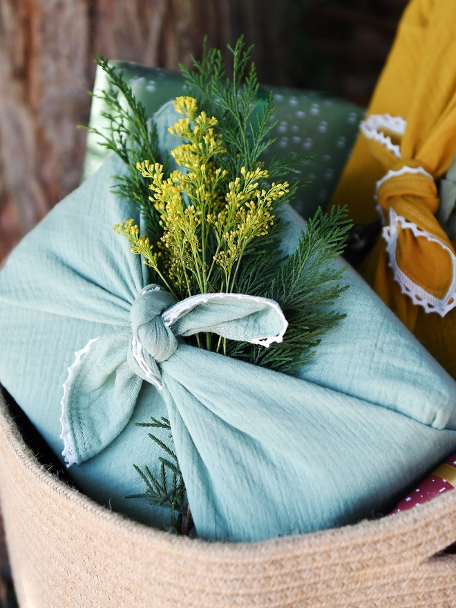 A gift wrapped in mint green fabric wrap, with an ivory lace trim, sits in a woven basket with yellow flowers tucked against it.