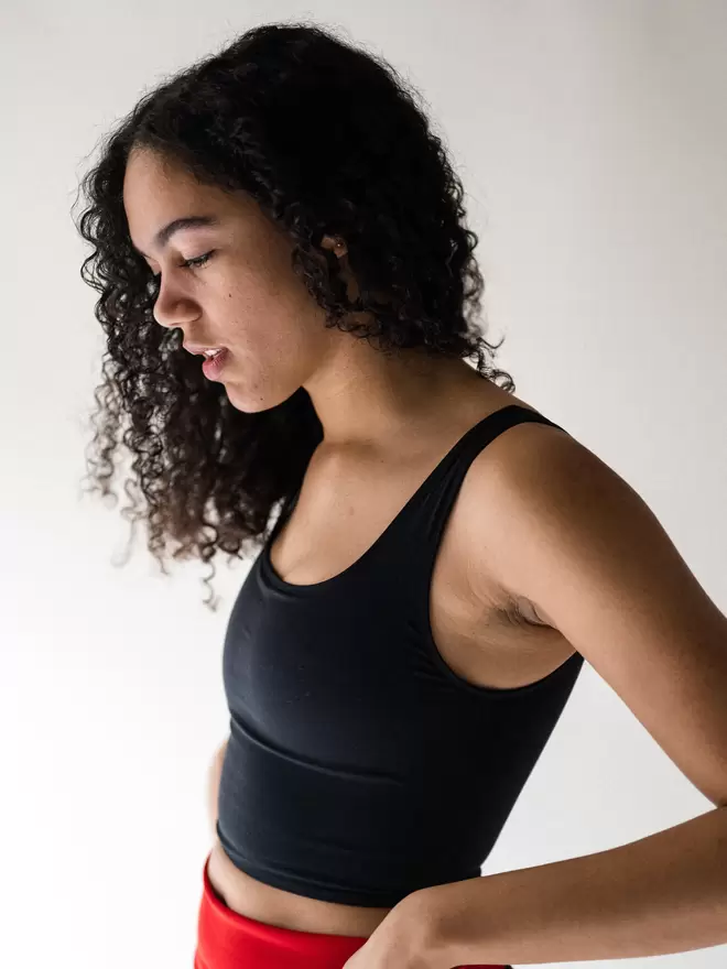 Side view of a lady with long black curly hair, looking down at the floor and hand on hip, wearing a Davy J Sustainable Waterwear black cropped swim top with square neckline and shoulder straps.