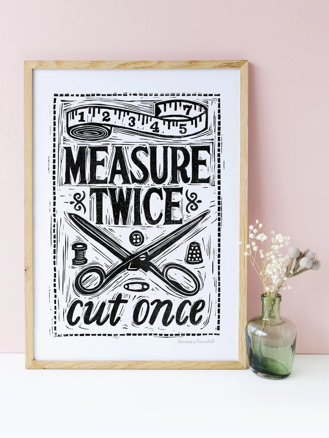 black and white sewing print in a wood frame with a green vase on a pink background