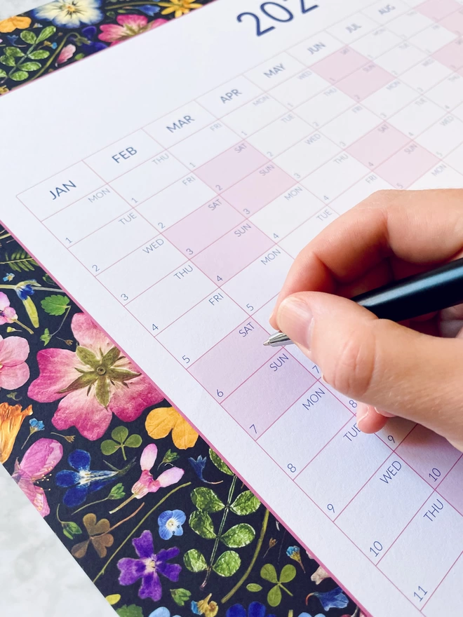 A hand holding a pen ready to write on a nature-inspired wall calendar with a pressed floral design