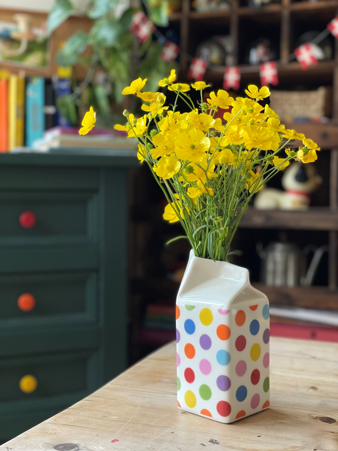 Colorful polka dot milk carton jug with yellow buttercup flowers on a wooden tabletop