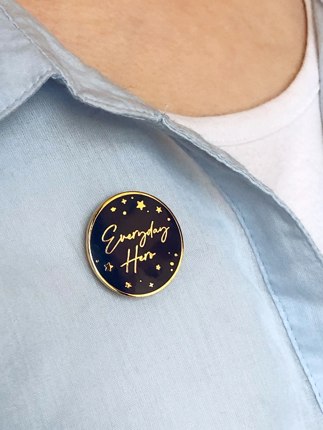 A navy blue and gold enamel pin badge with a gold star design and the words "Everyday Hero" is pinned to a blue shirt.
