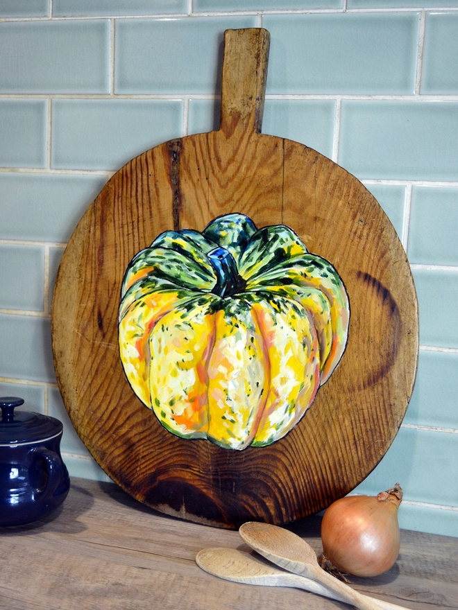Wooden chopping board with carnival squash painted design standing against a kitchen wall