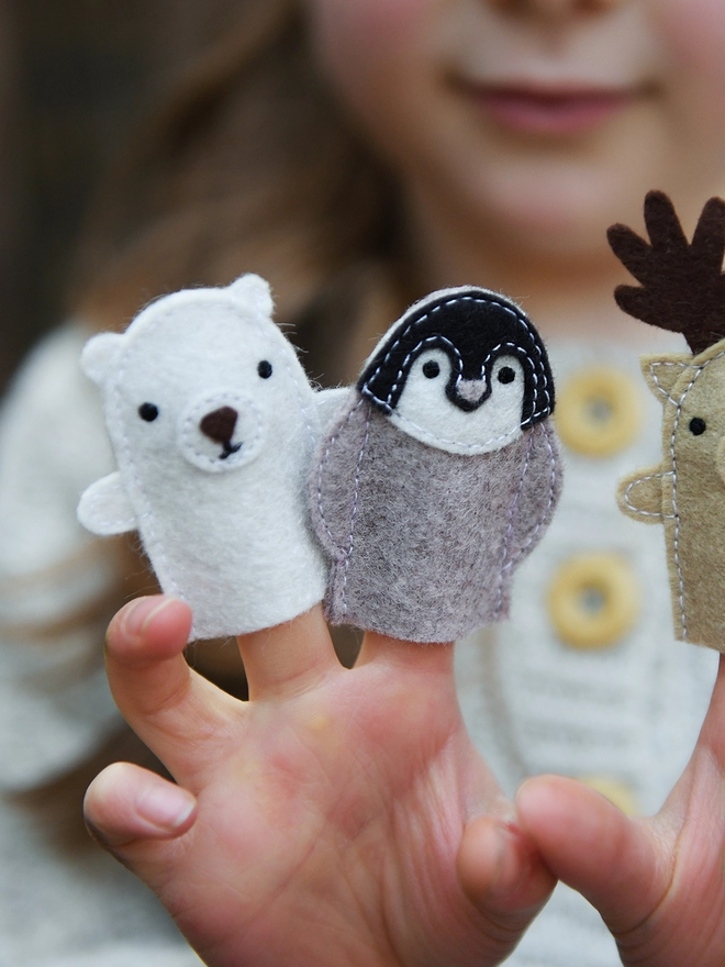 A young child holds three felt finger puppets on her fingers, a reindeer, a penguin, and a polar bear.