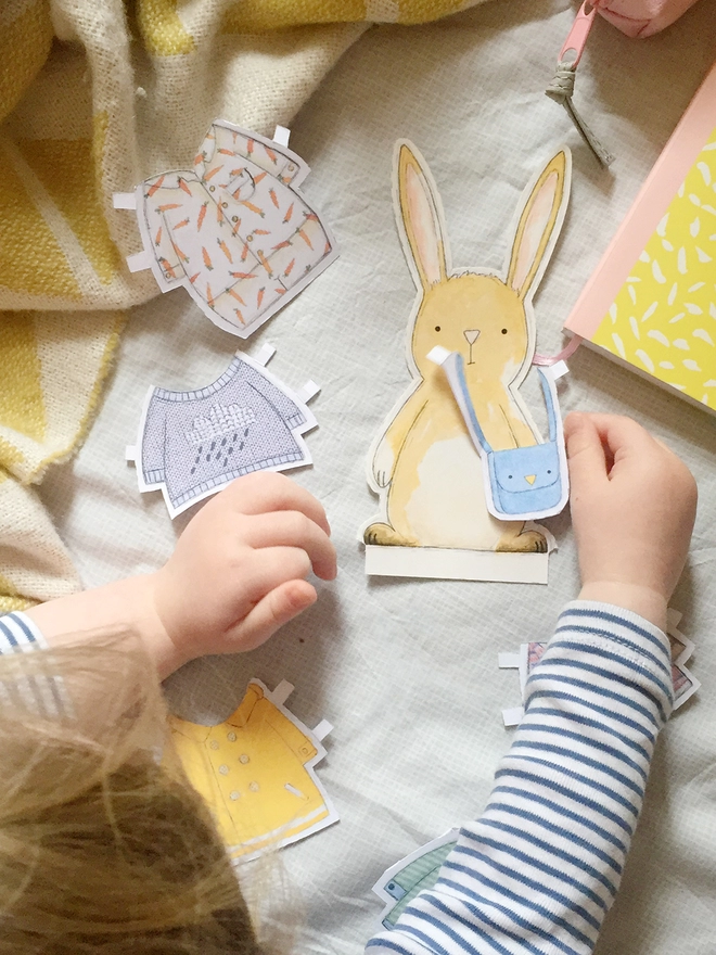 A young child is playing with a rabbit paper doll and dressing it in various paper outfits.