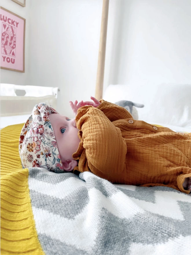 A baby with big blue eyes lies on its back on a double bed in a bright and neutral adult bedroom. The baby has its hands in its mouth and is lying on a grey and white chevron baby blanket with a mustard yellow trim. A print on the wall behind reads ‘Lucky You’.