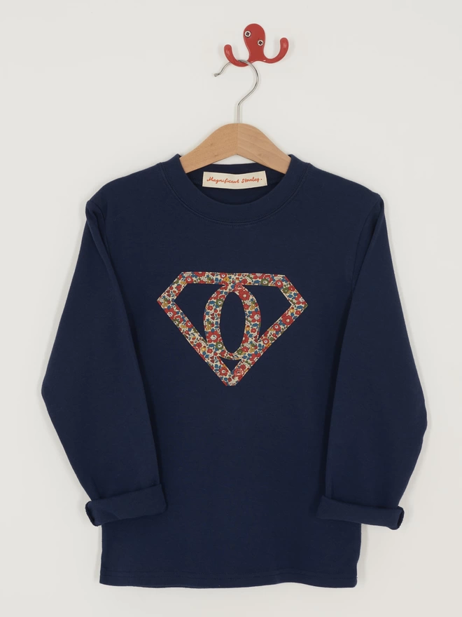 A navy long sleeve cotton t-shirt featuring the initial O inside a superhero motif. The motif is appliquéd in floral Liberty Print. T-shirt is hanging on a hanger.