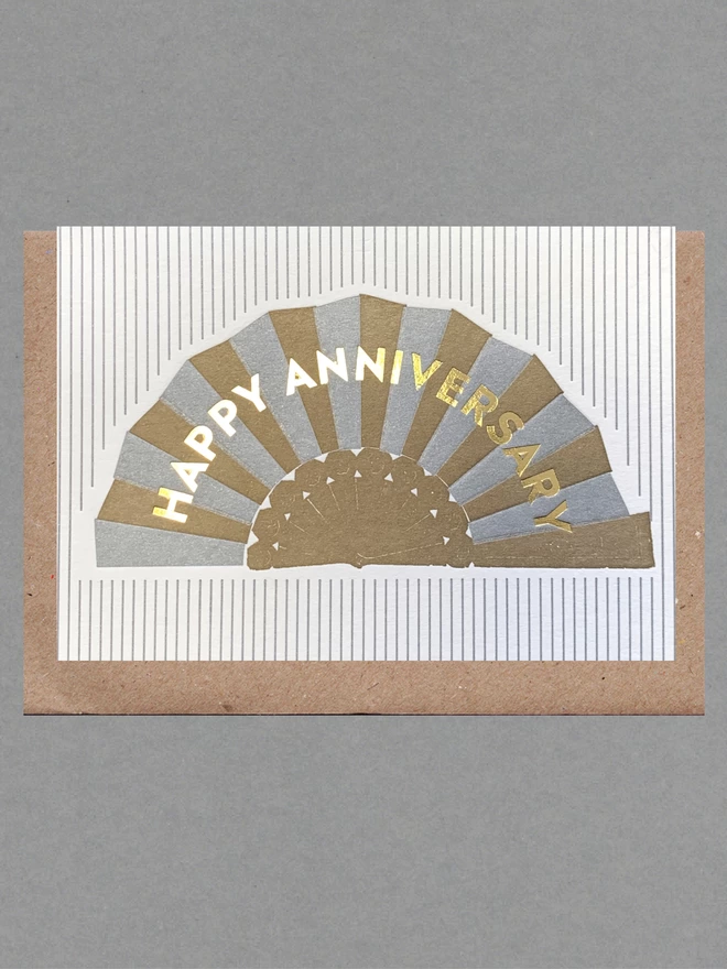 White card with silver and gold fan with gold text reading 'Happy Anniversary' on silver and white striped card with a brown envelope behind