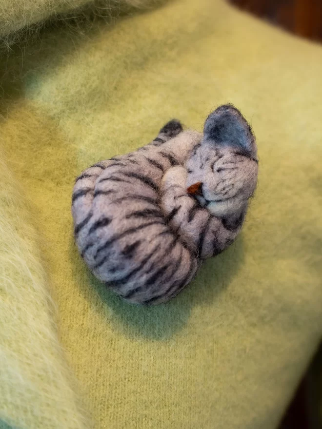 A needle-felted sleeping ginger silver cat brooch on a green fluffy jumper