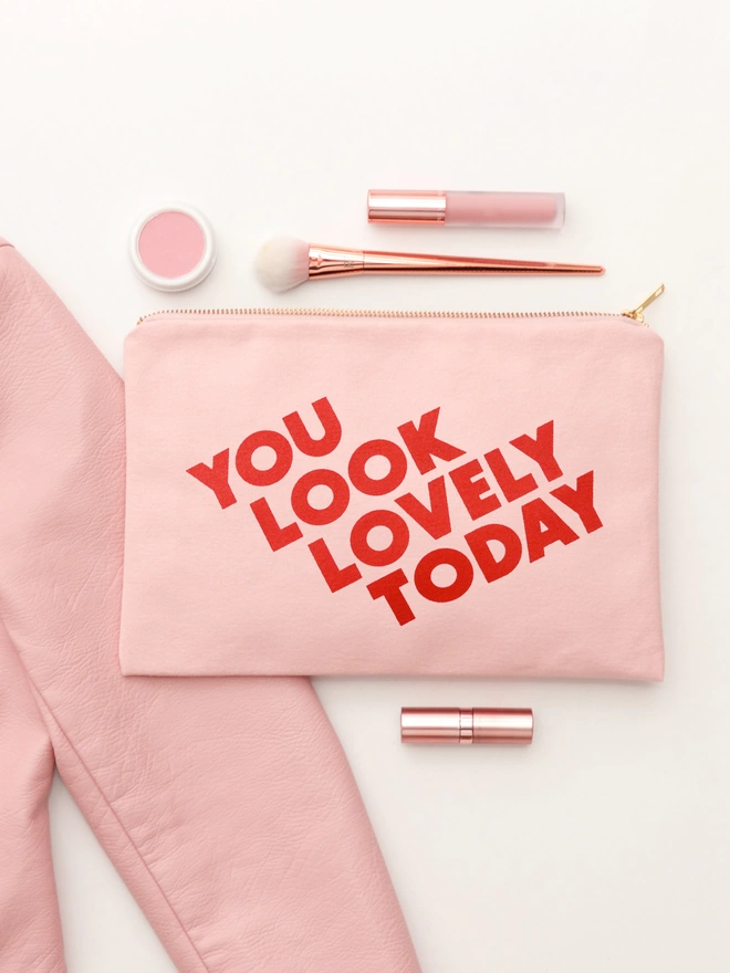 a blush pink canvas zip pouch with the words you look lovely today laying on a white backdrop with makeup surrounding