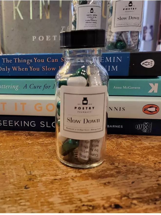 Glass pill bottle containing green and white Slow Down poetry pills printed on banana paper- displayed next to a pile of books on Poetry Pharmacy counter