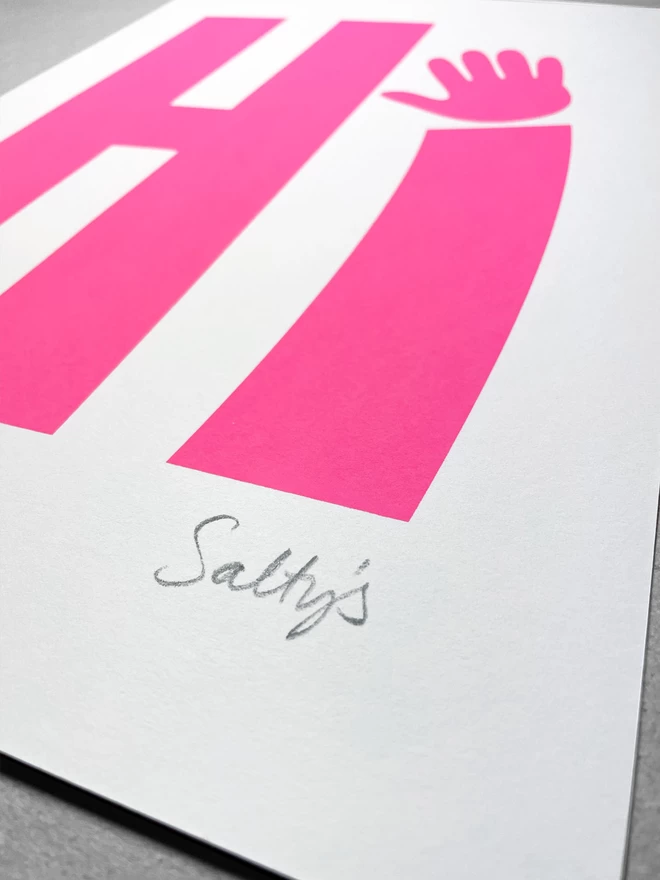 Close up of a neon pink printed Hi upon a white sheet on paper - sat on a grey background. CLose up shows the signature in the bottom right corner