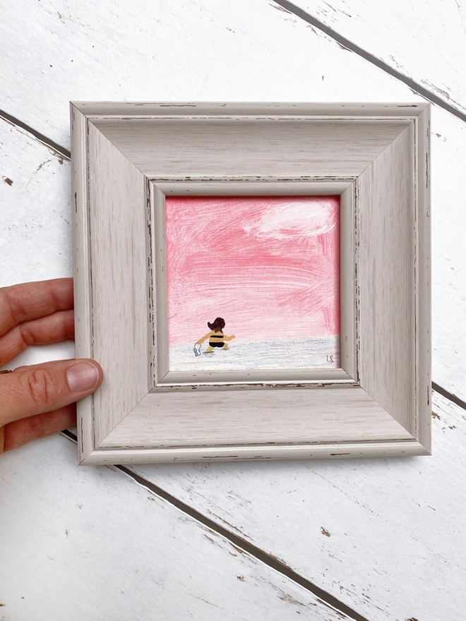 hand holding painting of sunset and swimmer in chunky pale grey frame