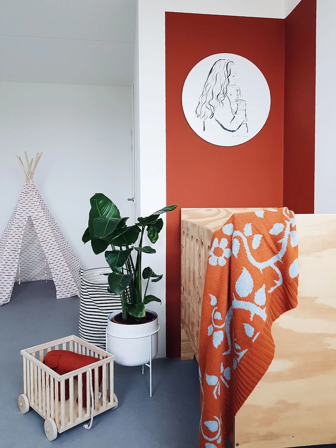View of a modern nursery showing the rust briar rose baby blanket draped over the end of a birch ply cot. A teepee tent can be seen in the background alongside a plant and a child's wooden pull along toy.
