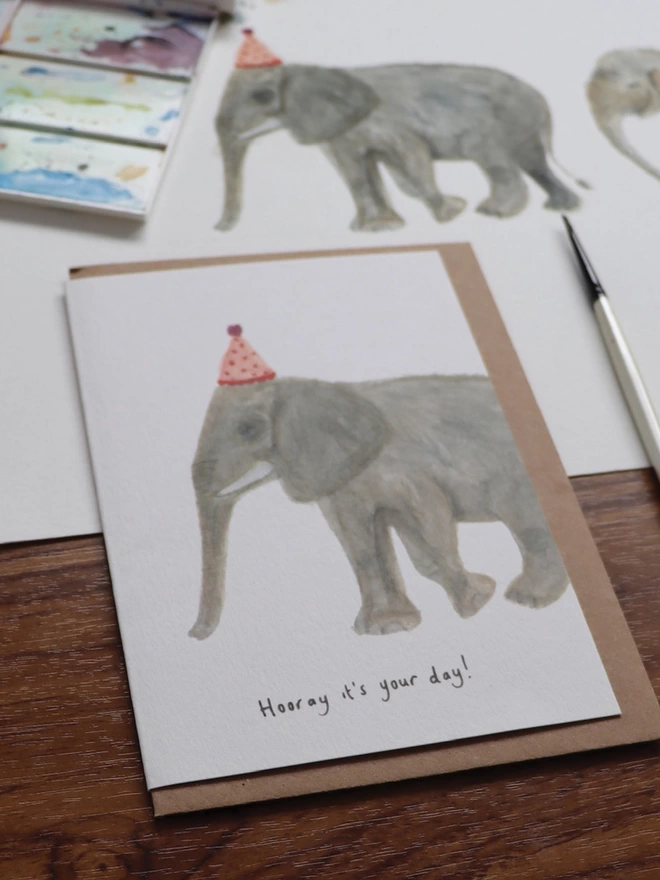Close Up Slightly Out Of Focus Shot Of The Elephant Greeting Card Sitting Along Side The Original Hand Painted Watercolour Illustration, Paintbrush and Palette
