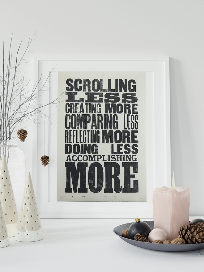 Scrolling Less - Letterpress Poster seen in a white frame with a candle and acorn in front