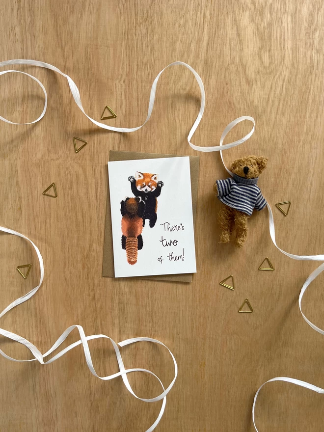 A greetings card featuring two baby red pandas stood up looking at each other with their arms in the air, next to the phrase “There’s two of them!”