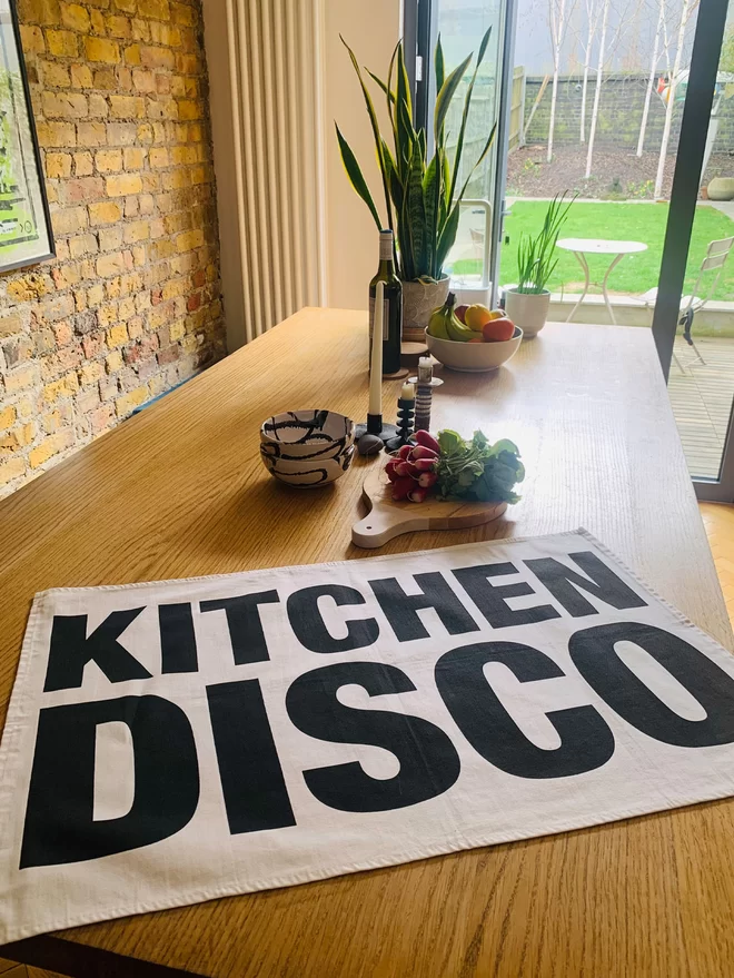 London Drying Kitchen Disco screen printed in black on white tea towel laid on table with chopping board, bowls , plant in background