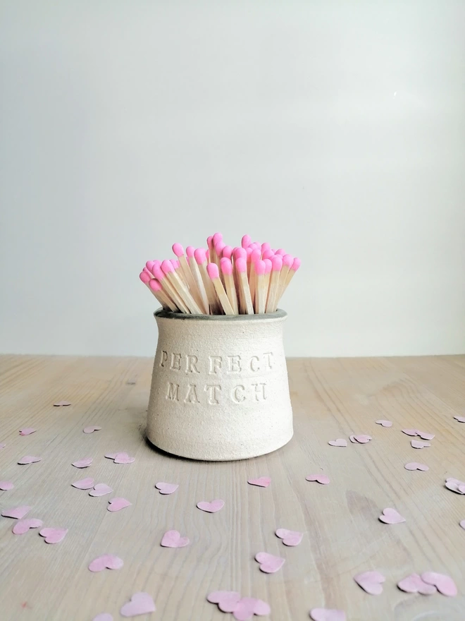 handmade match pot on wooden surface surrounded by pink paper hearts. Pink tip matches inside the pot. Grey glaze showing on rim of match pot. Perfect Match wording on the outside of the pot