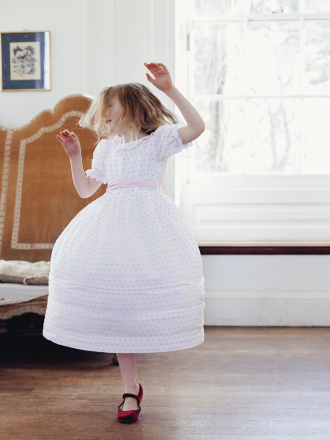 A little girl dances in a pink and white swiss dot dress with a pink sash