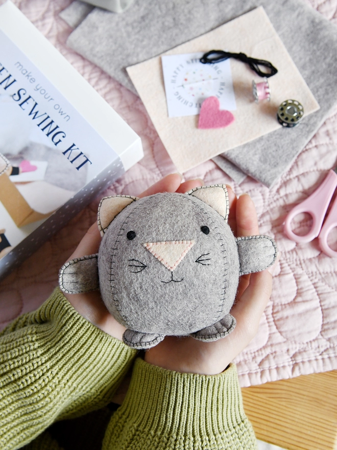 A handmade felt kitten toy is being held in two hands above a desk covered in craft kit components.