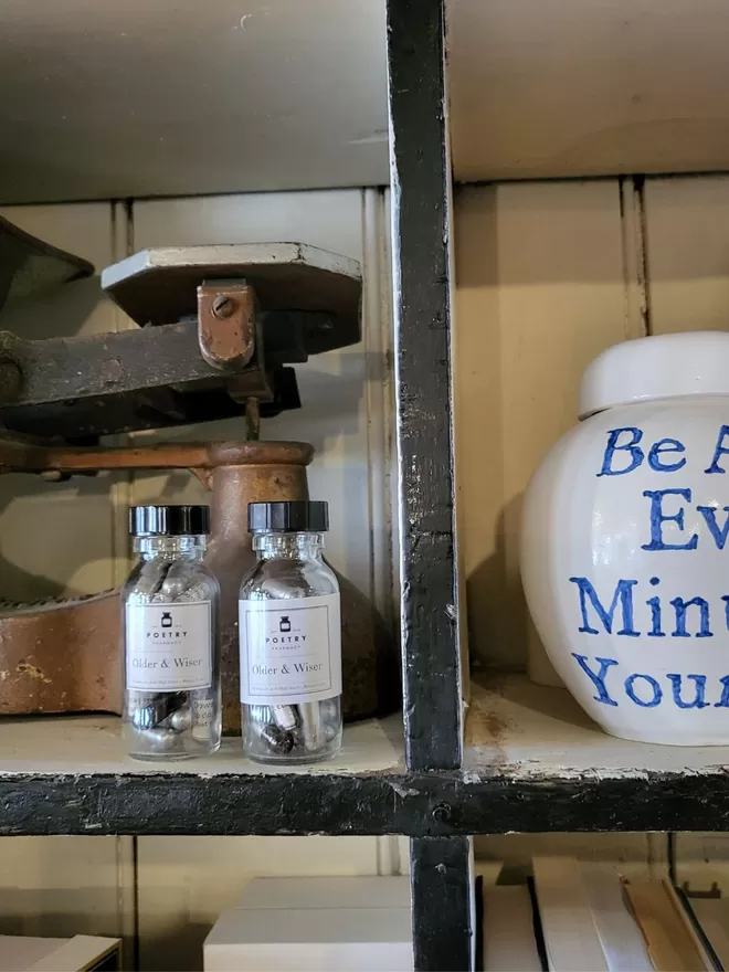 Glass pill bottles containing silver, black and clear Older & Wiser poetry pills printed on banana paper on Poetry Pharmacy shelves