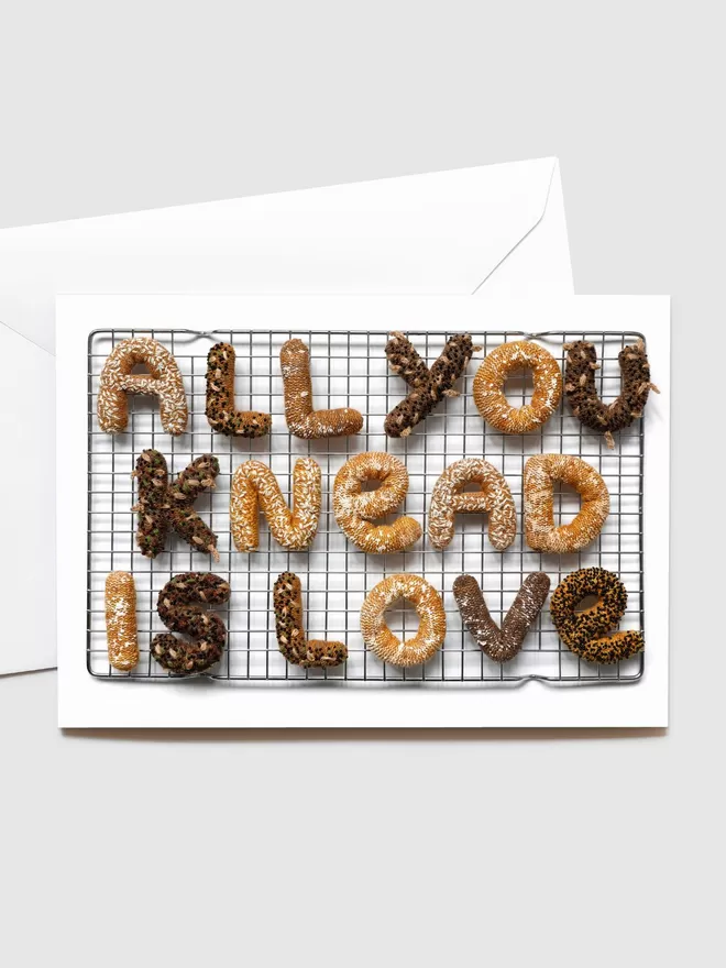 Kate Jenkins crotched and hand embroidered bread spelling out 'All You Knead Is Love' on top of a baking tray. The white envelope for the Blank Card is seen behind.