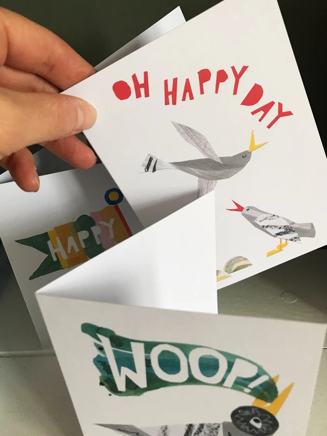 Set of three illustrated greetings cards by Esther Kent showing colourful lettering and stylised birds. A hand pics out a card that reads 'Oh Happy Day' in red.
