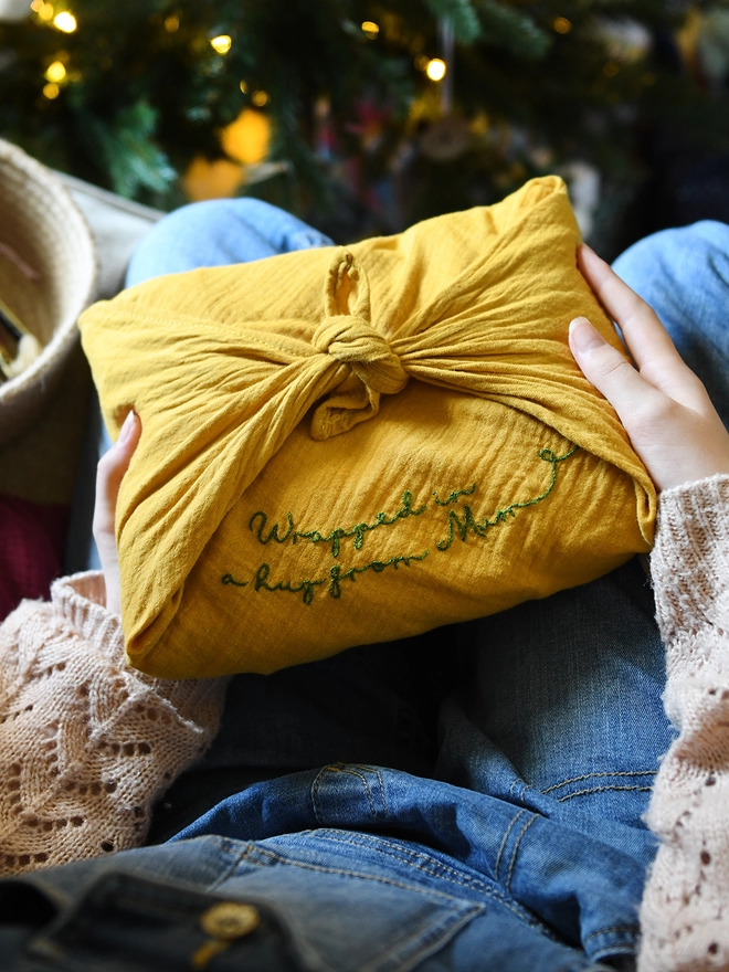 A gift wrapped in a mustard yellow cotton fabric wrap that has been embroidered with a special message. 