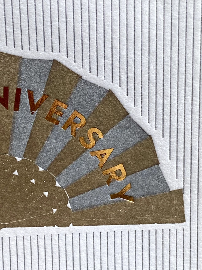 Close up of White card with silver and gold fan with gold text reading 'Happy Anniversary' on silver and white striped card.