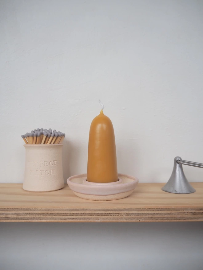 Shelf containing stoneware candle holder in pale pink, next to it a match pot with matches and candle snuffer