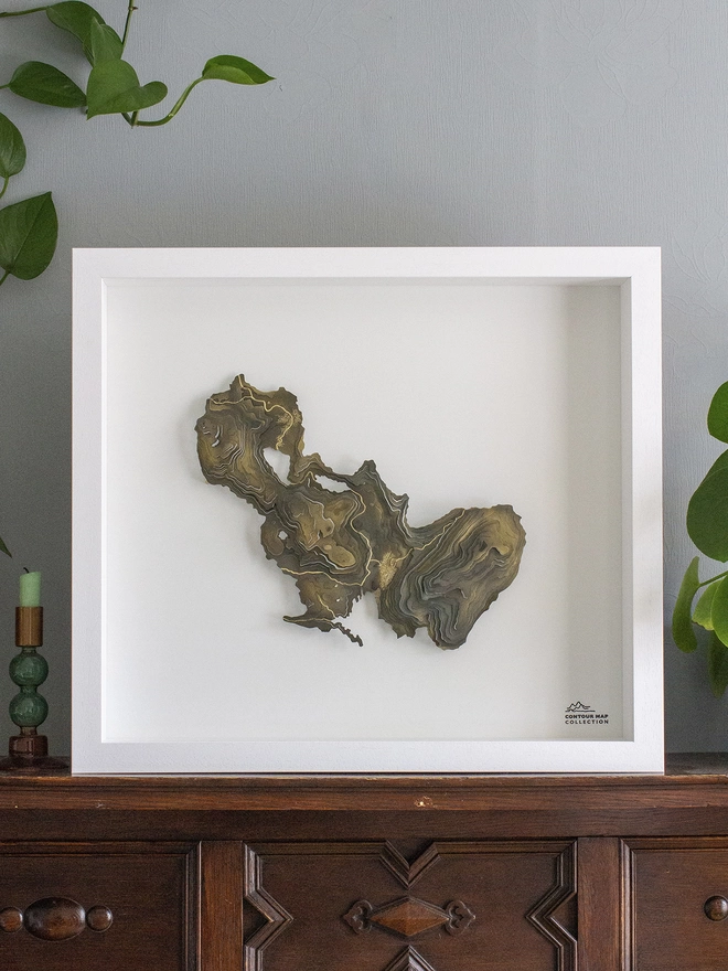 A bespoke metal contour map wall piece with a very irregular outline, mounted on white in a white frame.