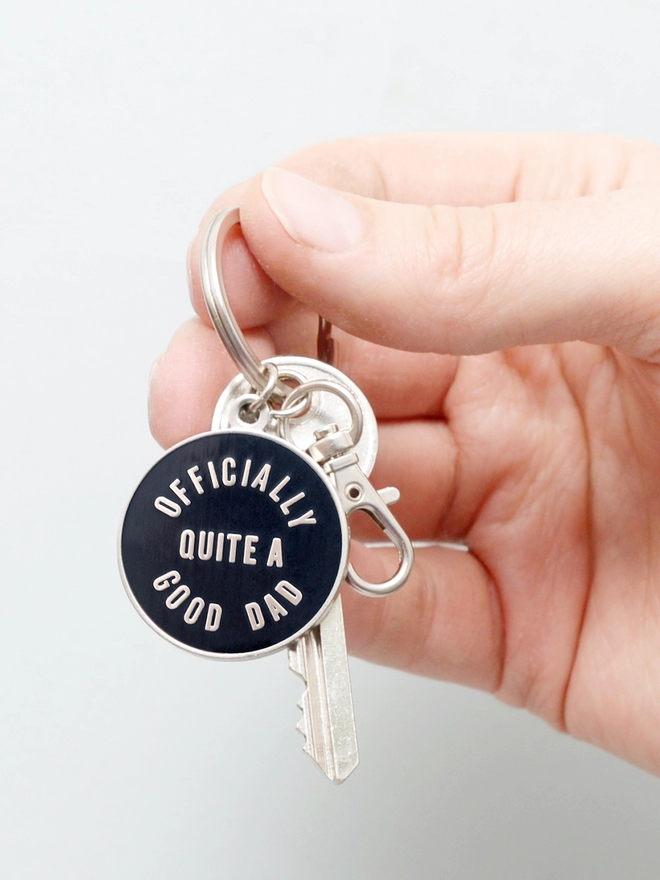 Hand holding out a Black and silver enamel keyring with 'Officially Quite a Good Dad' design