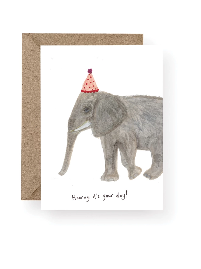 Elephant Birthday Card, Card Has a White Base With Hand Painted Elephant Wearing A Party Hat.  Sitting On A Recycled Brown Kraft Envelope.  There Is Black Handwritten Text Underneath The Elephant Which Reads ‘ Hooray it’s your day’