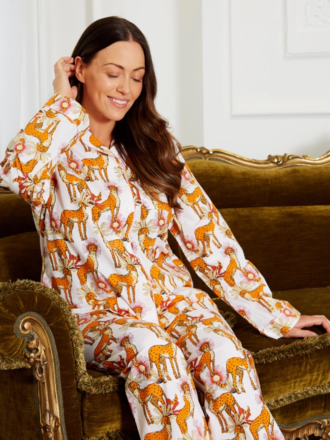 Model puts her hand behind her ear sitting on a velvet green couch wearing a pair of white based classic pyjamas with an orange cheetah and pink flower print
