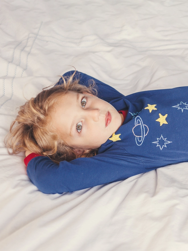 A boy in navy and red pjs with a space scene lies on a bed
