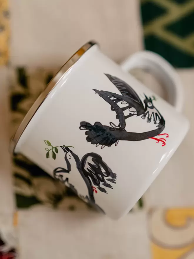 White enamel mug decorated with black inky dove drawings lives on a patterned quilt