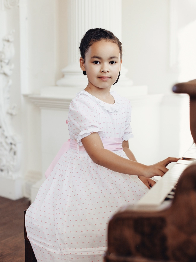 A little girl sits at a piano in a pink and white swiss dot dress with a pink sash