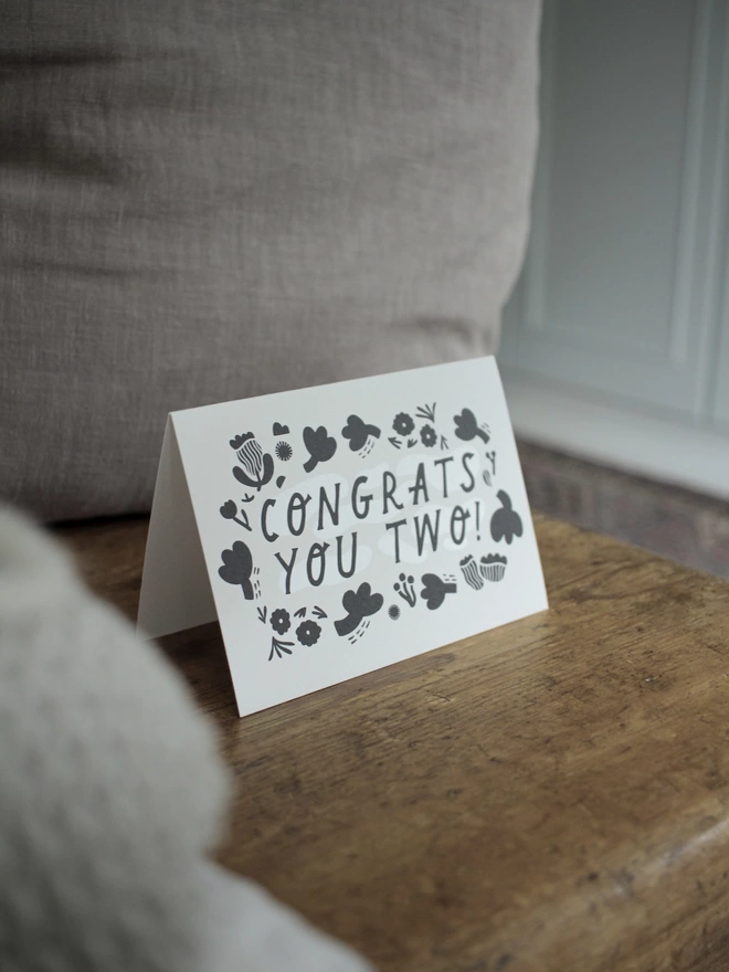 Black and white greeting card with illustration and the words Congrats you two written on it stood up on wooden surface with a grey cushion behind it 
