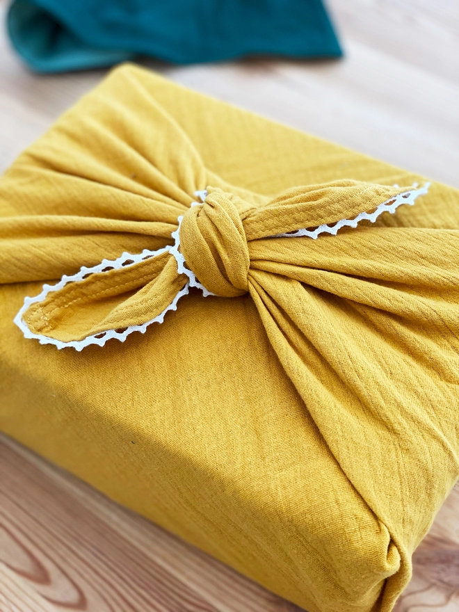 A gift wrapped in mustard yellow cotton fabric wrap with an ivory lace trim is on a wooden desk.